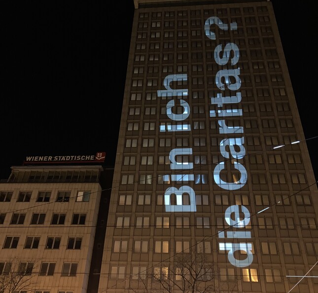 Nachts projizierter Schriftzug „Bin ich die Caritas?“ auf hohem Gebäude