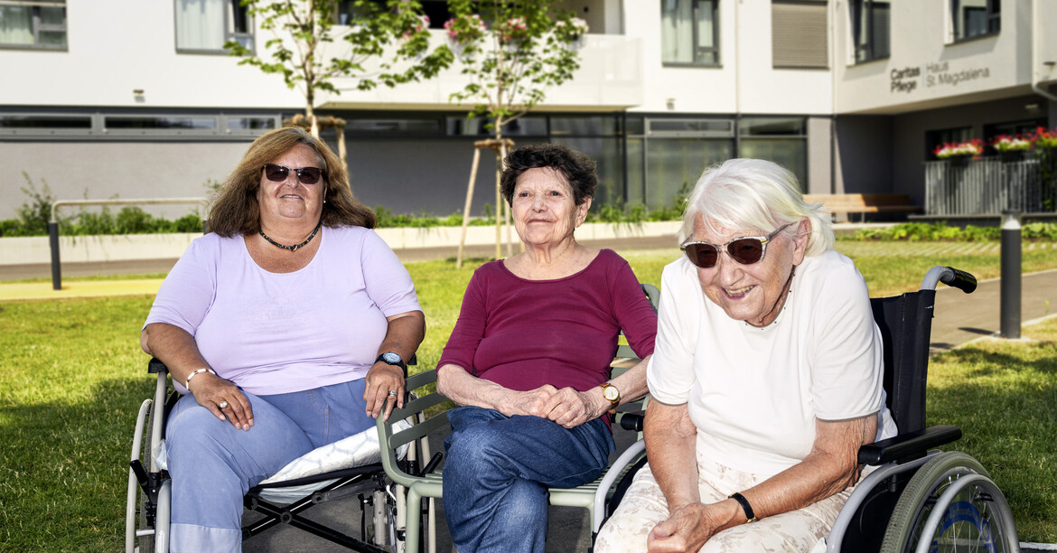 Drei ältere Frauen sitzen auf ihren Rollatoren bzw. Rollstühlen nebeneinander, sie lächeln in die Kamera. Im Hintergrund ist ein Pflegewohnhaus zu erkennen.