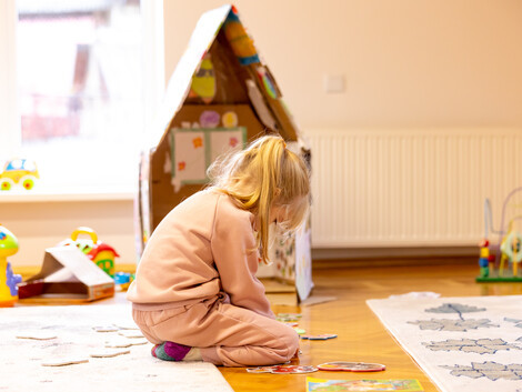 Ein Kind sitzt am Boden und spielt, im Hintergrund ist ein Spielzeughaus und Spielzeug erkennbar.