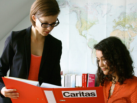 Zwei junge, erwachsene Frauen sind in einem Büro. Beide sind in rot gekleidet. Eine zeigt der anderen einen roten Ordner, auf dem das CARITAS Logo zu sehen ist. Eine der beiden Frauen trägt eine Brille.